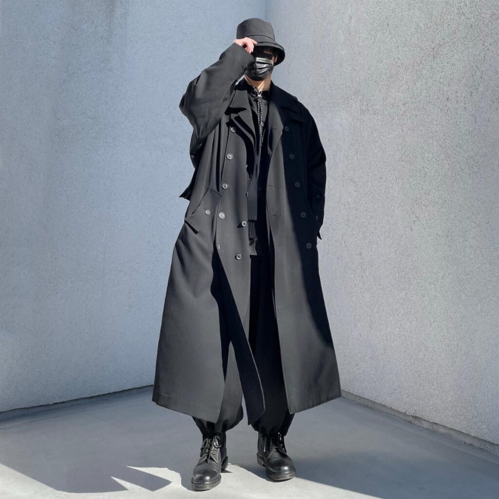 A person wearing an all-black long trench coat, trousers, boots, and a bucket hat.