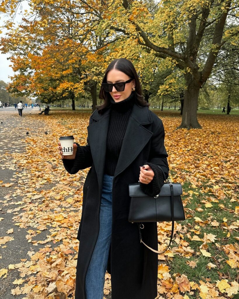 Woman in a long black coat and sunglasses walking through an autumn park with fallen leaves.
