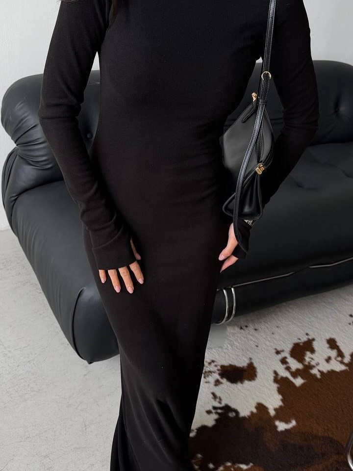 A person wearing a long-sleeved black maxi dress standing near a black leather sofa.