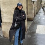 Woman wearing a long black trench coat, blue jeans, and a navy baseball cap on a city street.