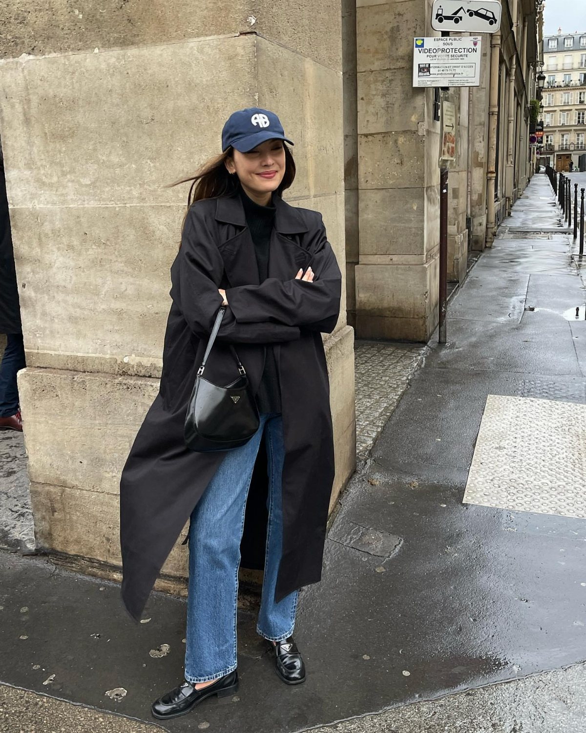 Woman wearing a long black trench coat, blue jeans, and a navy baseball cap on a city street.