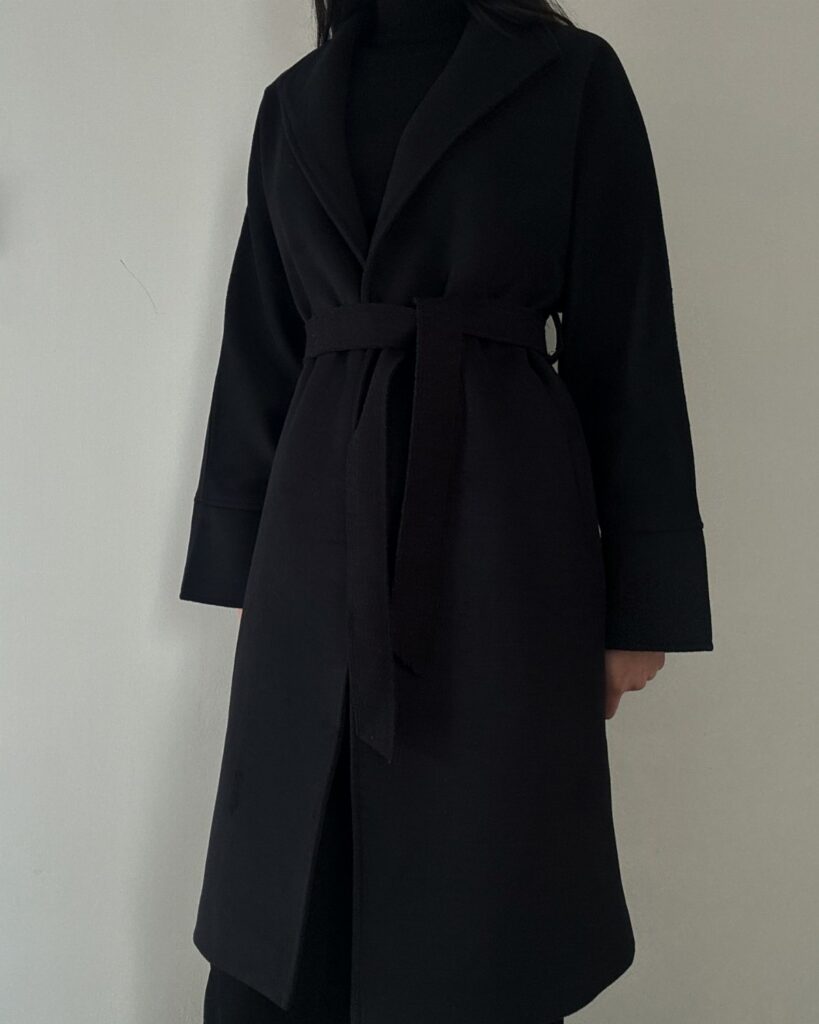 A person wearing a long black wrap coat with a belted waist against a white wall.