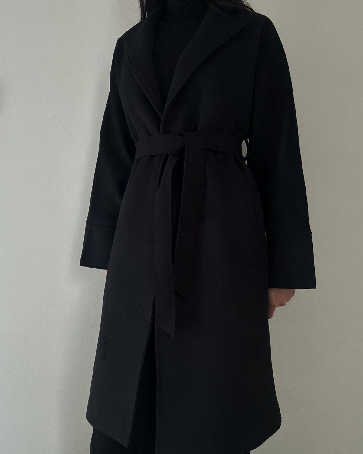A person wearing a long black wrap coat with a belted waist against a white wall.