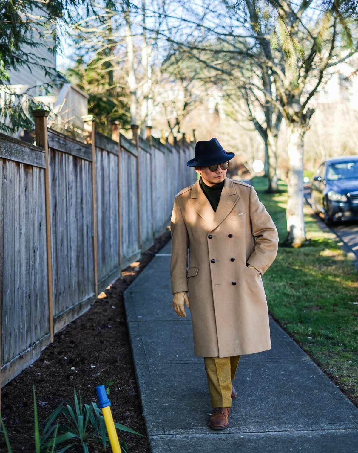A person in a tan double-breasted overcoat and dark hat walking on a sidewalk.