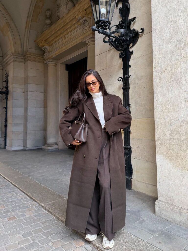 Woman in a long brown wool coat, white turtleneck, and brown trousers standing outdoors.