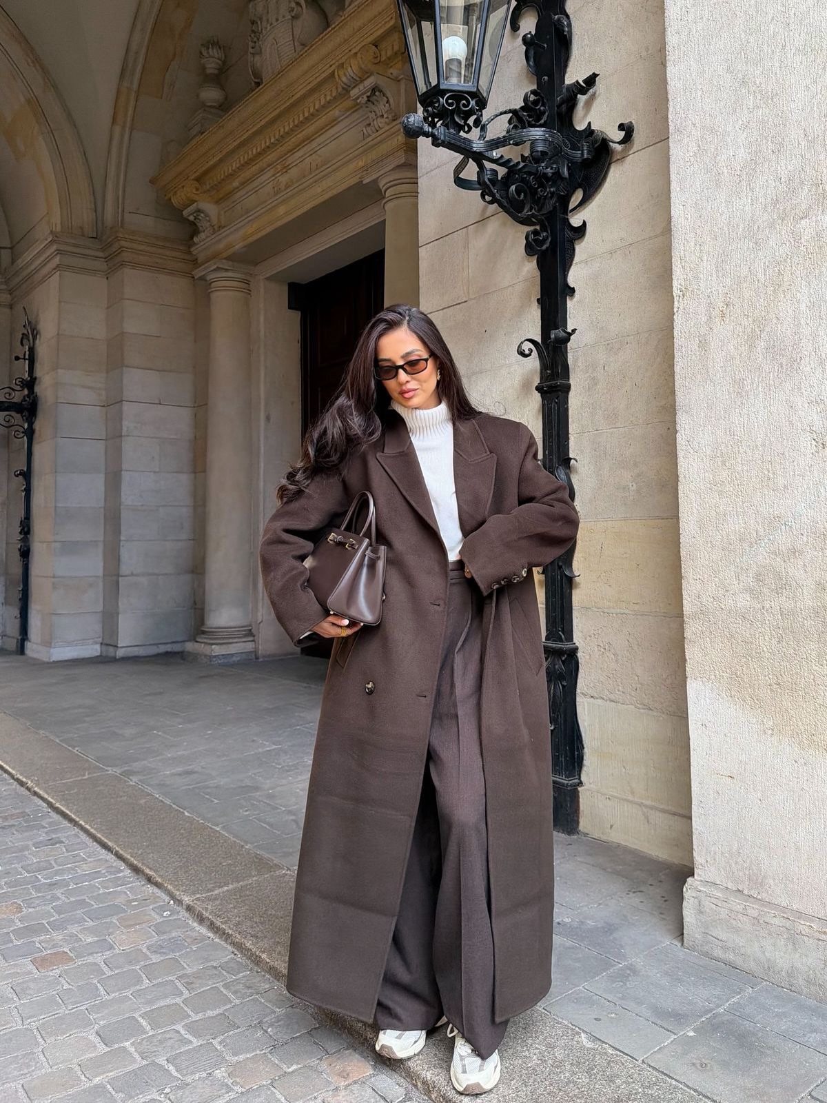 Woman in a long brown wool coat, white turtleneck, and brown trousers standing outdoors.