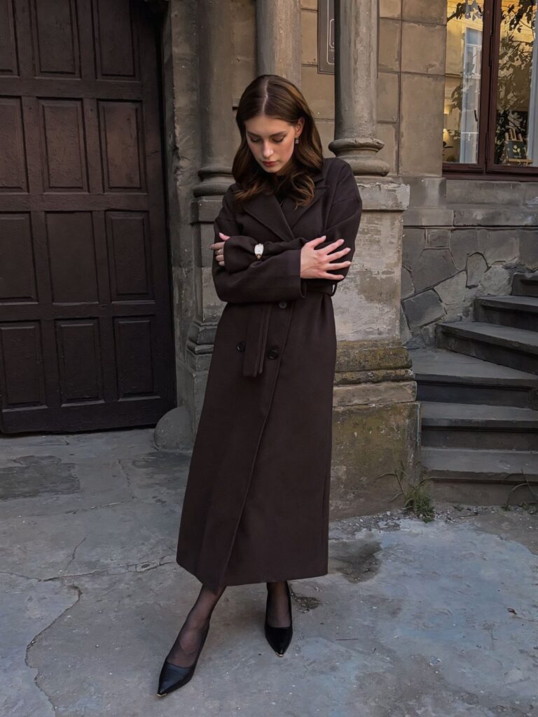 Woman wearing a long dark brown wool coat, black tights, and black heels standing outdoors.