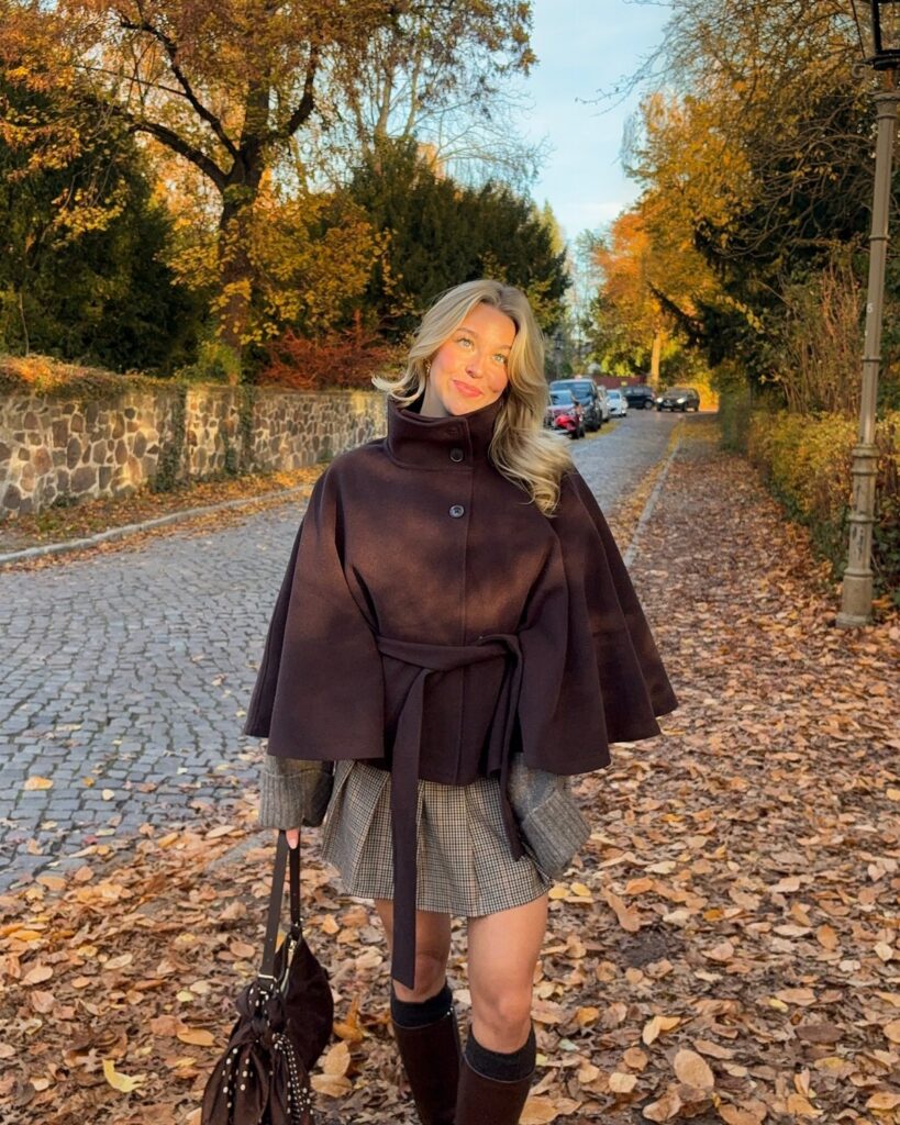 Woman wearing a dark brown belted cape coat and plaid skirt on a leaf-covered path.