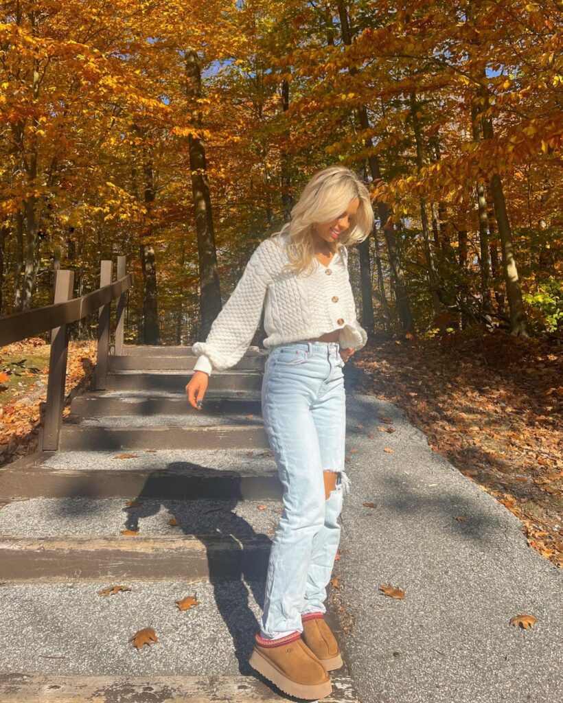 Blonde woman in a white knit sweater and light wash jeans walking through autumn trees.