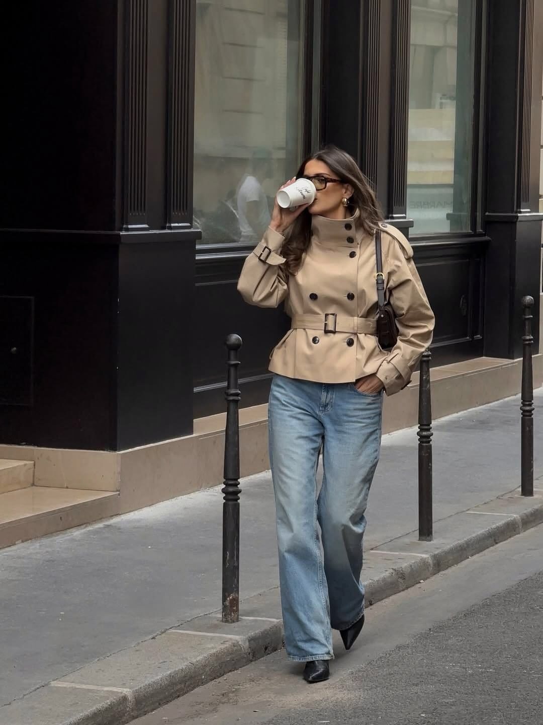 Woman in a cropped beige trench coat and wide-leg blue jeans walking on a city street.