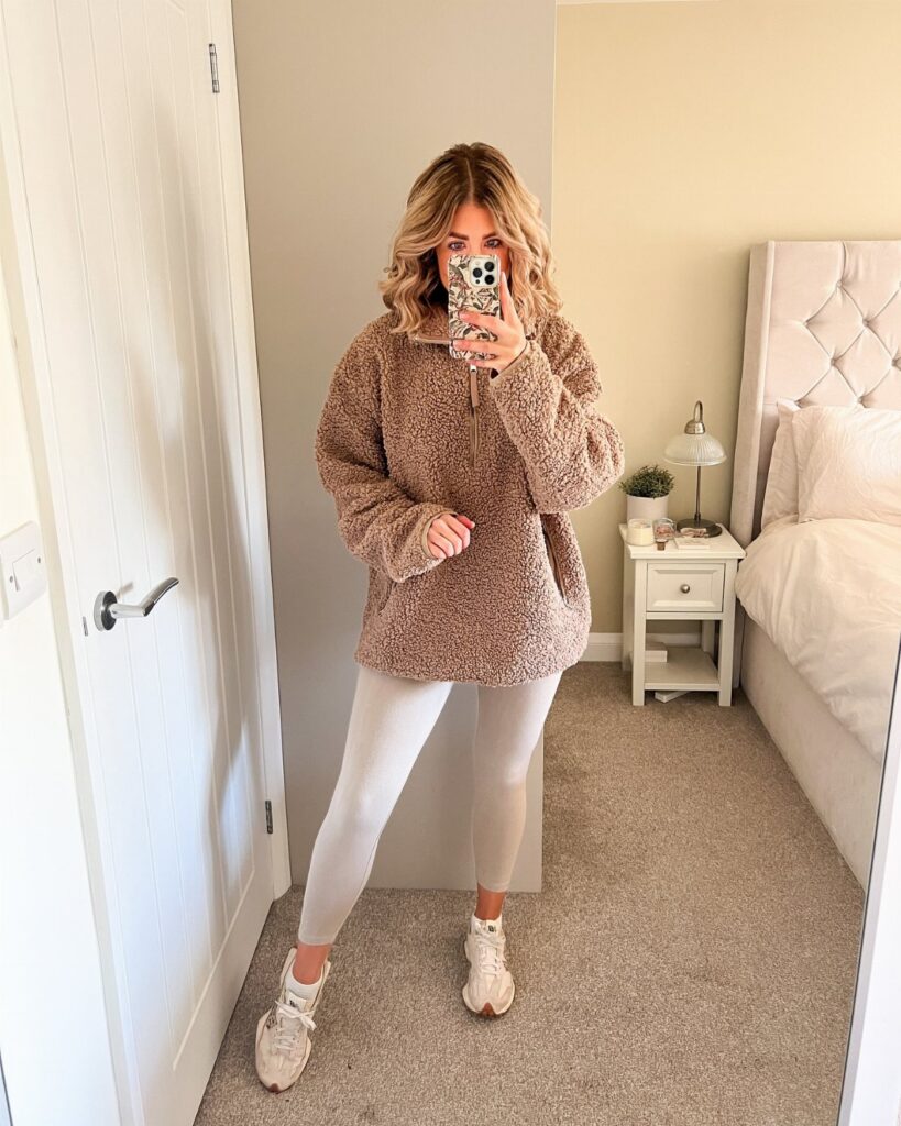 Woman in a fuzzy brown pullover sweater and beige leggings posing for a mirror selfie.
