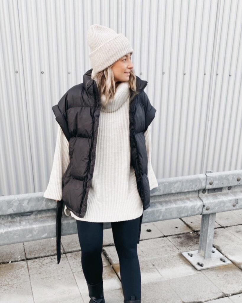 Woman wearing a cream oversized turtleneck sweater, black leggings, a black puffer vest, and a cream beanie.