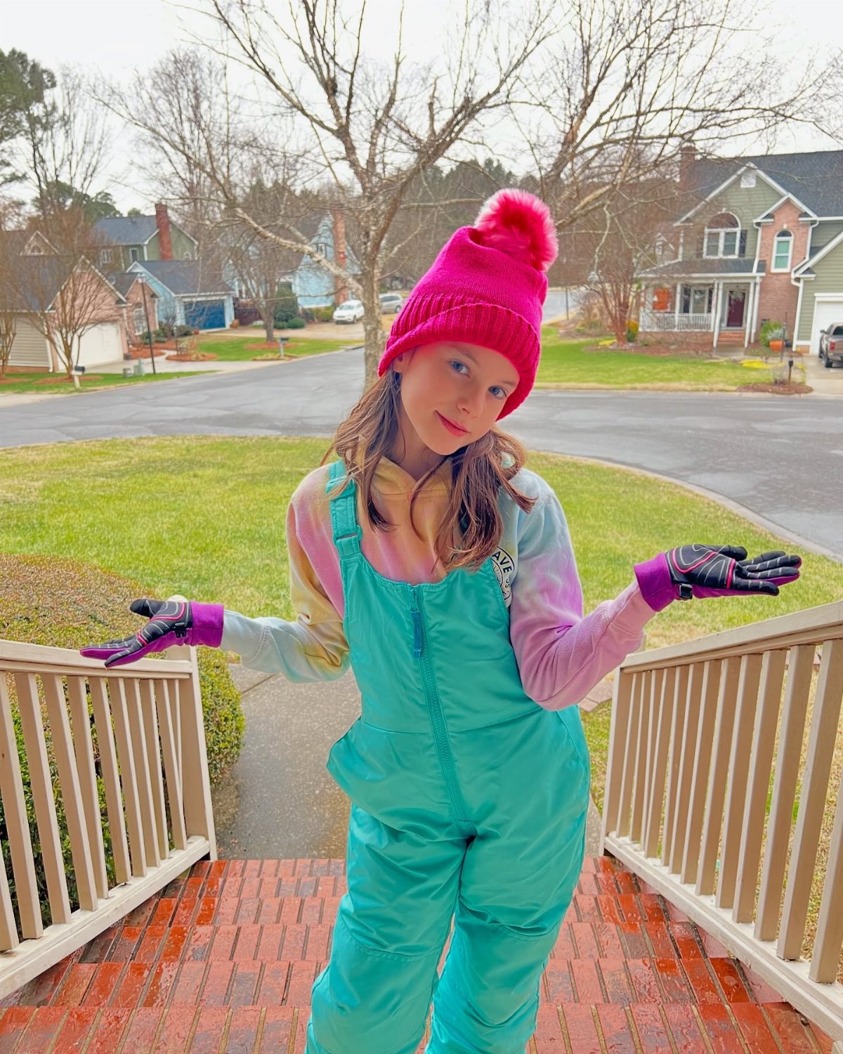A young girl in teal snow overalls and a pink pom-pom beanie posing on a porch.