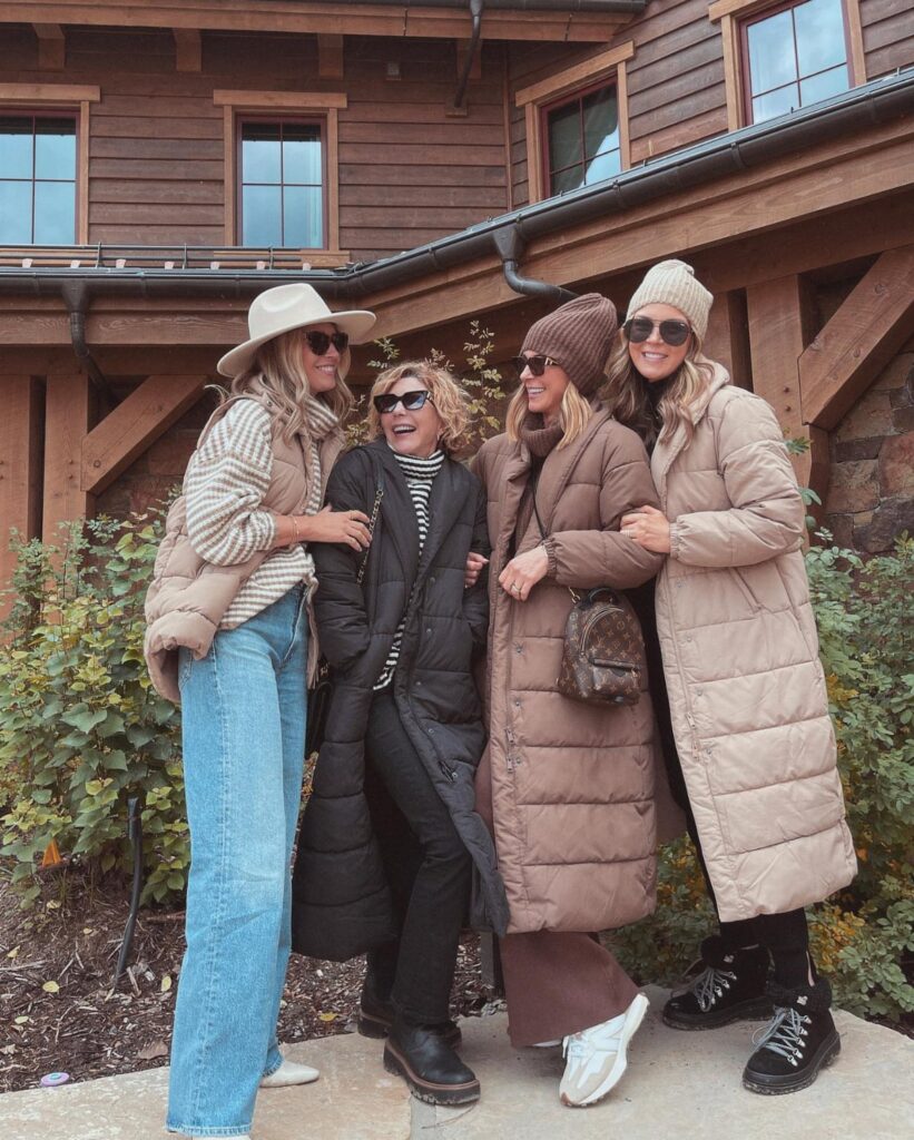 Four women posing together wearing stylish winter coats, knit hats, and warm layered clothing.