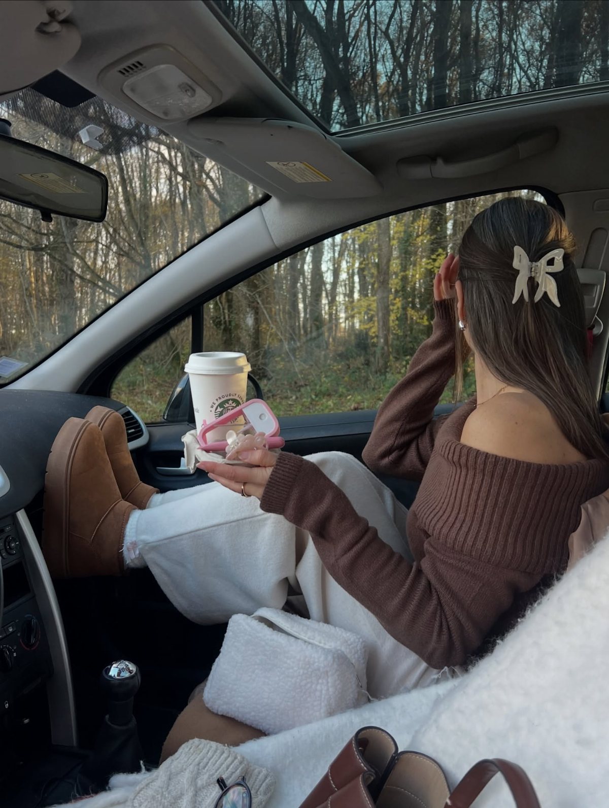Woman in a brown ribbed sweater and white pants sitting in a car looking at trees.