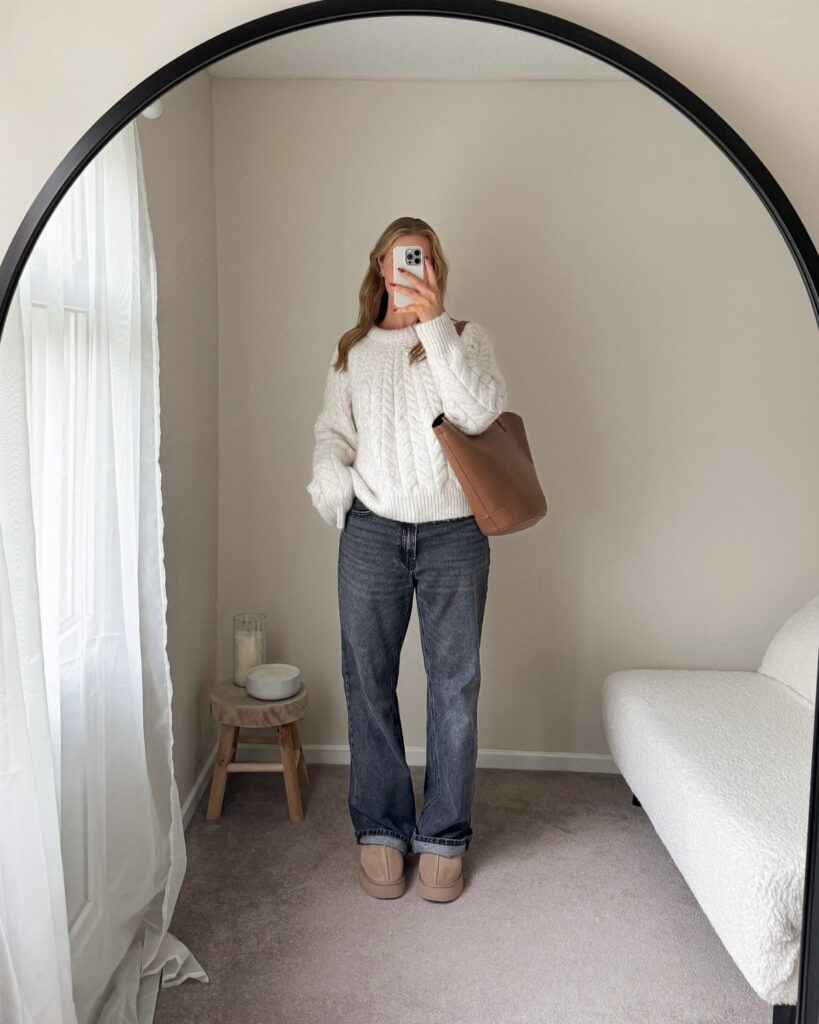 Woman in a white knit sweater and dark jeans posing for a mirror selfie.