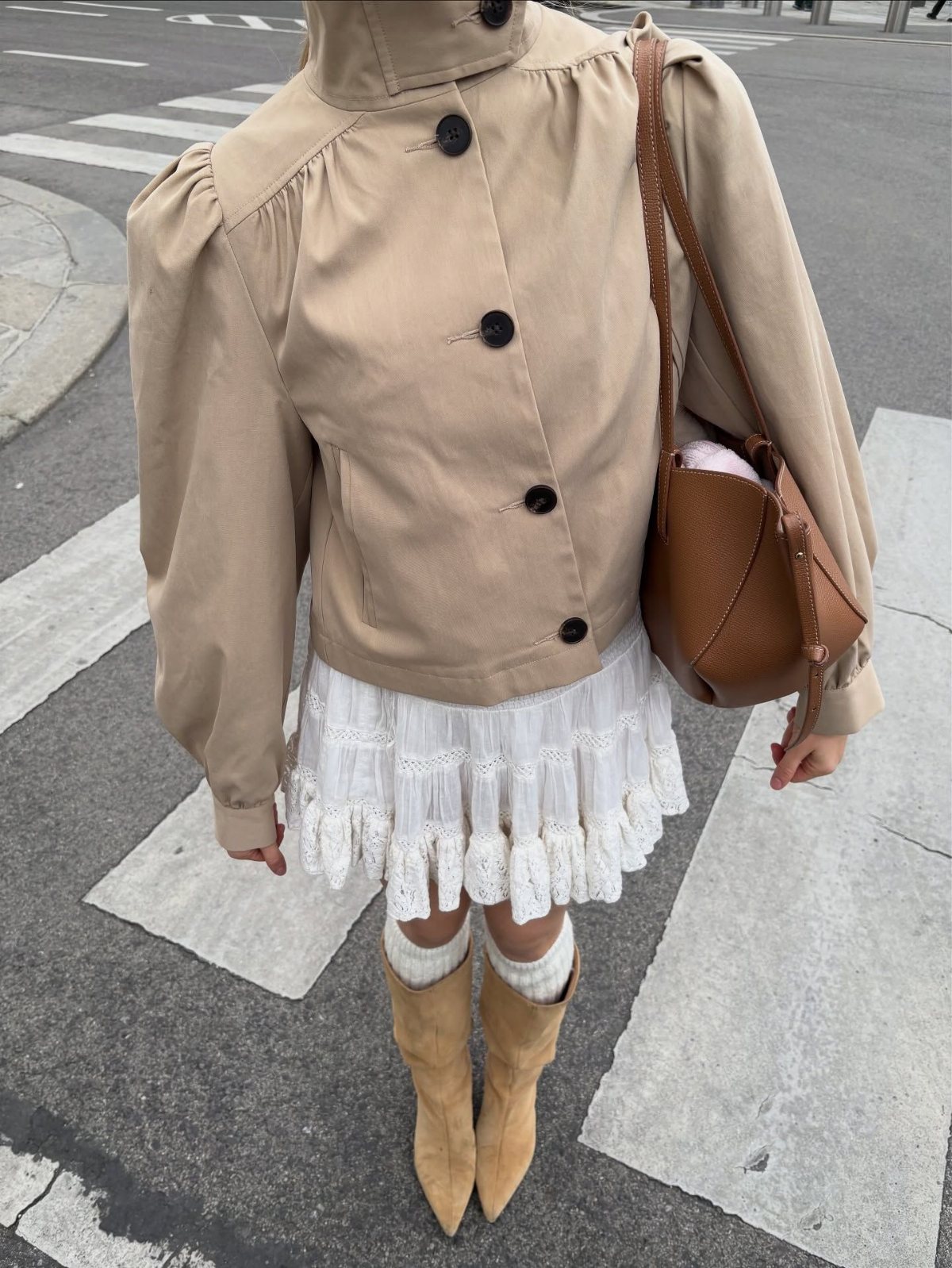 A person wearing a beige short trench coat, white lace skirt, and tan suede boots.
