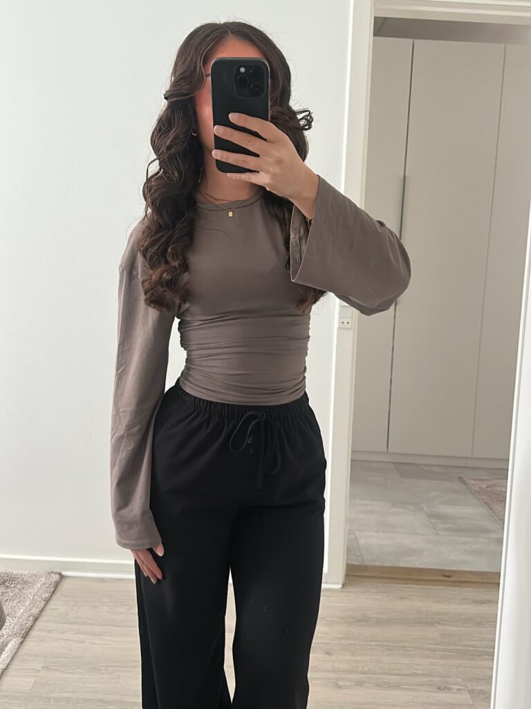 Woman in a taupe long-sleeved top and black trousers taking a mirror selfie.