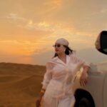 Woman in white desert attire leaning against a vehicle during a golden sunset.