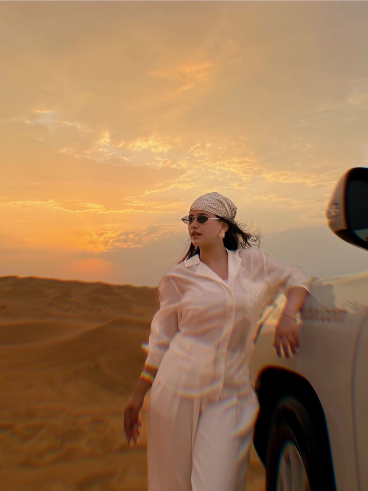 Woman in white desert attire leaning against a vehicle during a golden sunset.