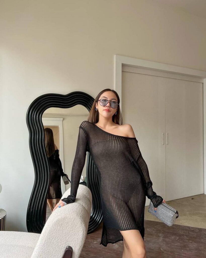 Woman in a black sheer knit off-the-shoulder mini dress posing in a room.