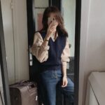 Woman in a beige button-down shirt and navy sweater vest posing for a mirror selfie.