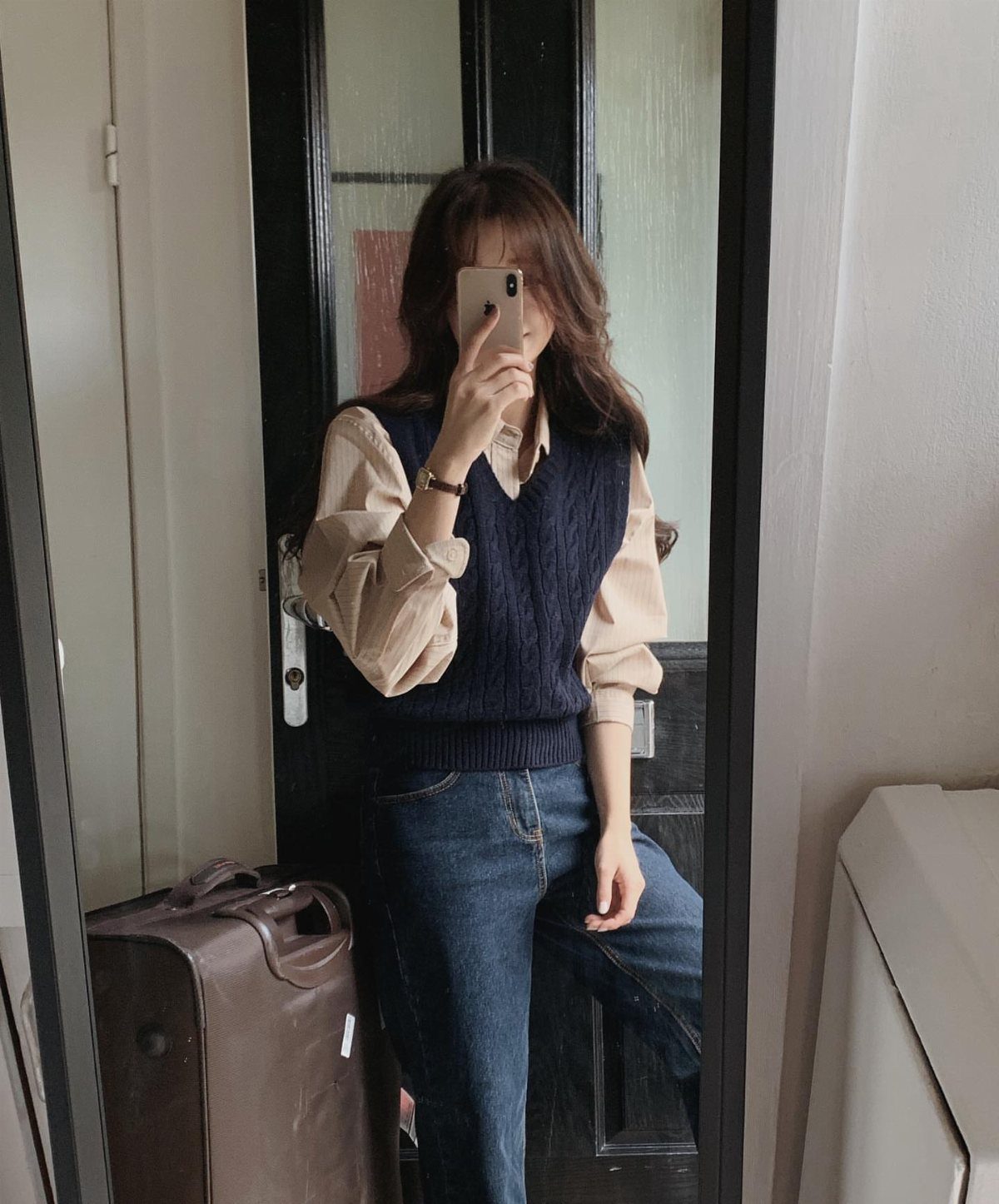 Woman in a beige button-down shirt and navy sweater vest posing for a mirror selfie.