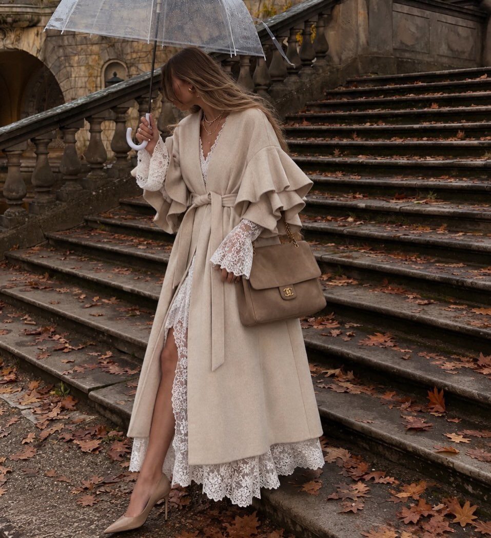 Woman in a beige ruffle sleeve coat and lace dress standing on autumn stairs.
