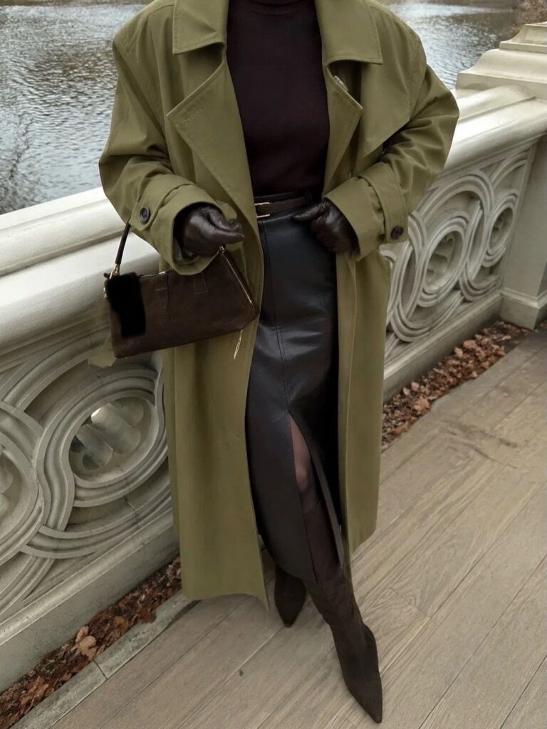 A person wearing an olive green trench coat, dark brown leather skirt, and dark gloves standing by a stone railing.