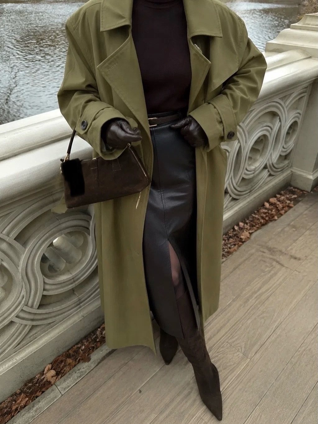 A person wearing an olive green trench coat, dark brown leather skirt, and dark gloves standing by a stone railing.