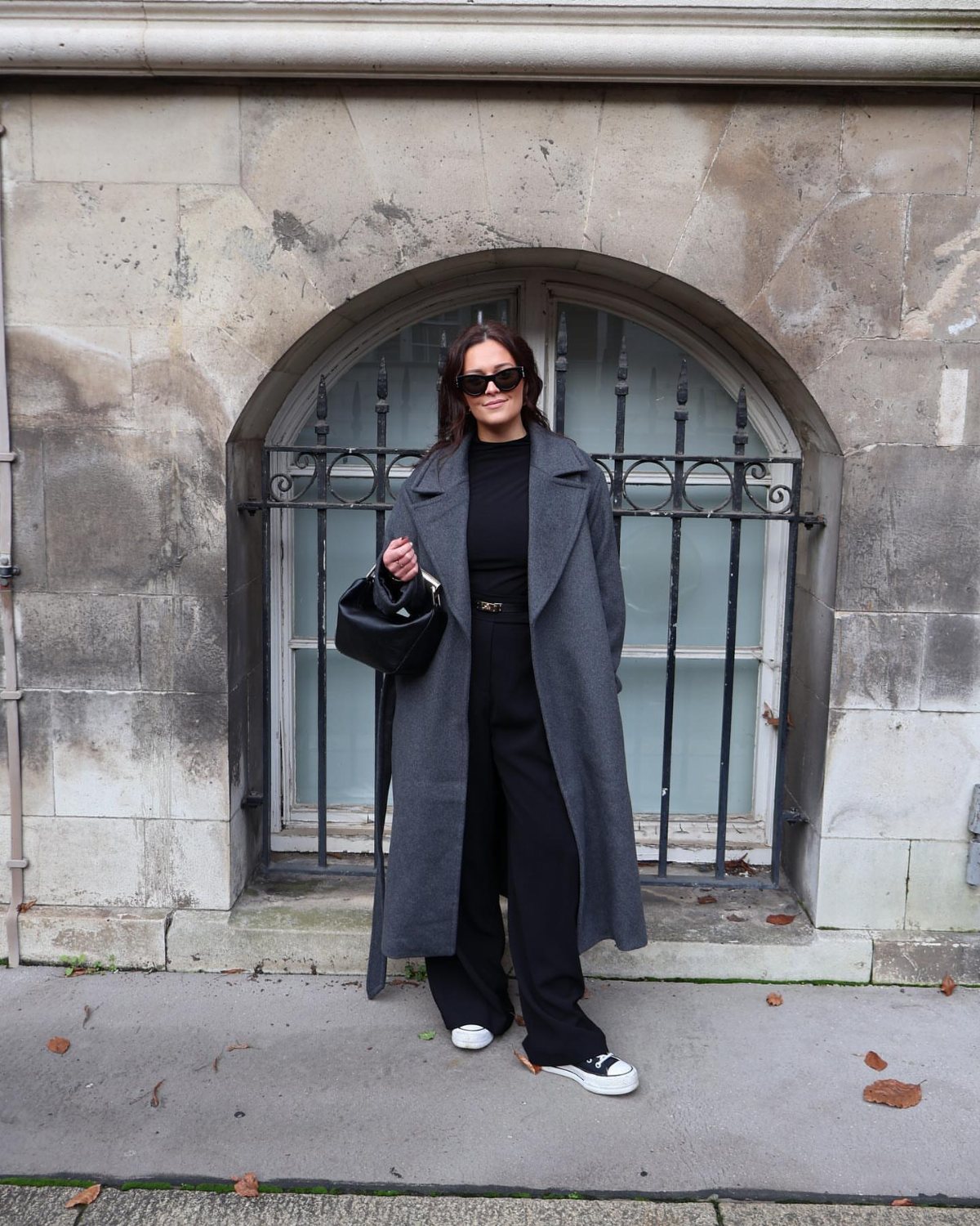 Woman in a long grey coat, black top, and wide-leg trousers standing by an arched window.