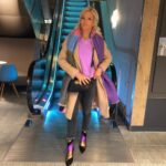 Blonde woman posing in grey jeans, a purple top, and a long camel coat.