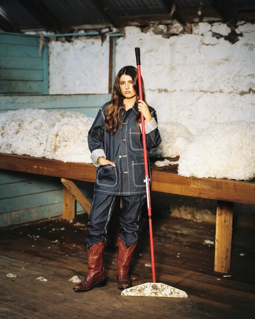 Woman in dark denim jacket and matching trousers holding a red broom in a barn.