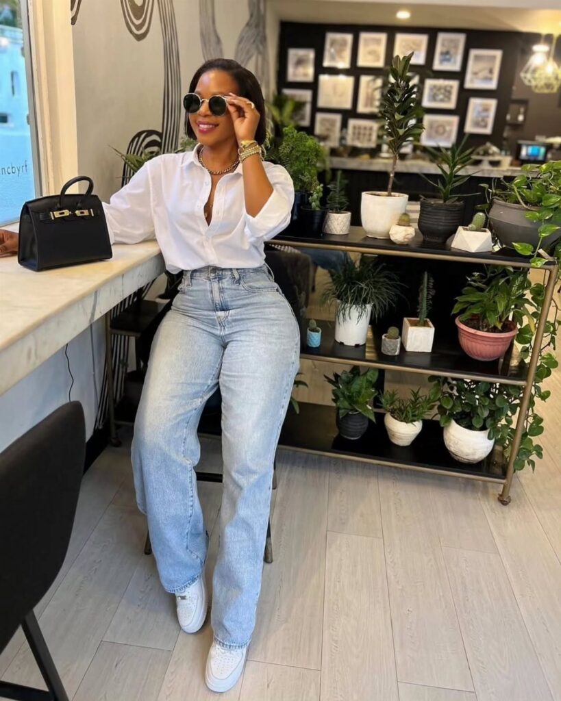 A woman wearing a white shirt and light blue wide-leg jeans standing in a room with plants.