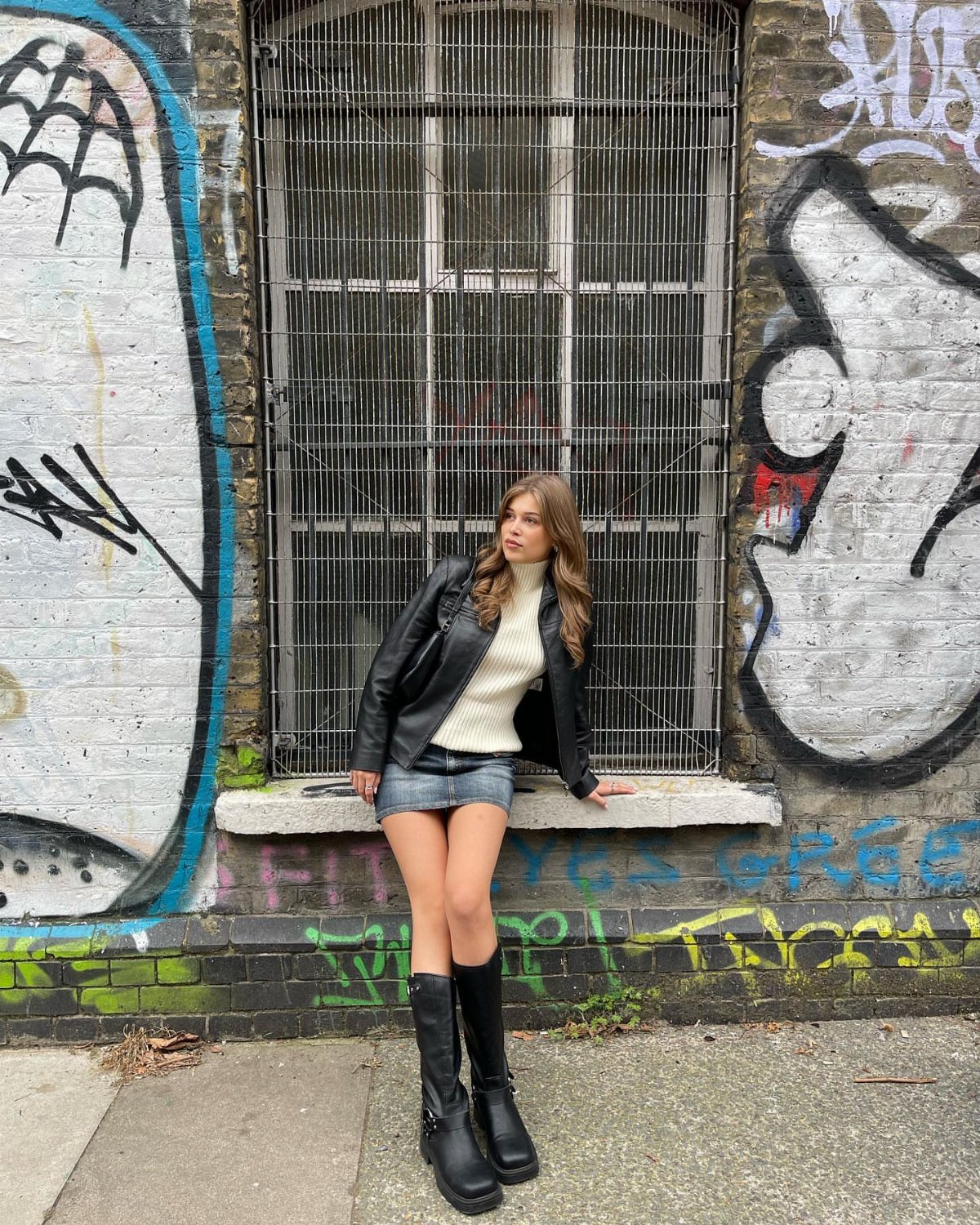 A woman wearing a black leather jacket, white turtleneck, denim skirt, and black boots posing against a graffiti wall.