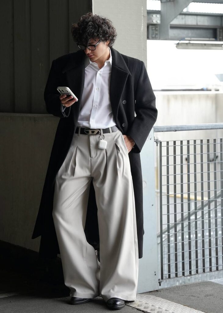A man in a long black coat, white shirt, and beige trousers looking at his phone.
