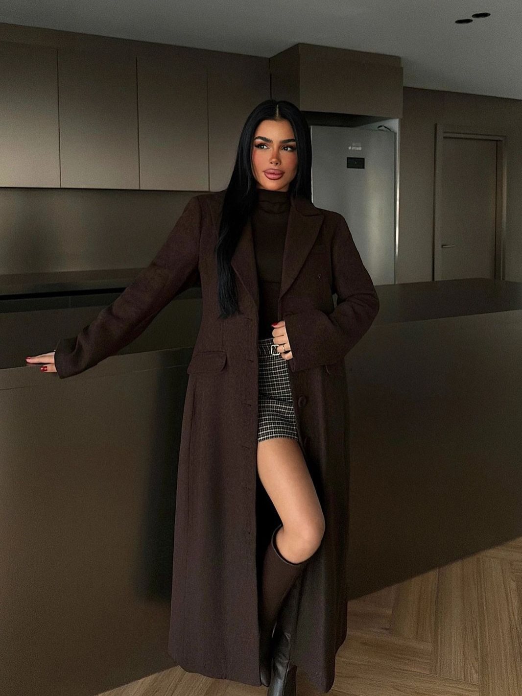 Woman posing in a long brown coat, plaid skirt, and black boots in a kitchen.