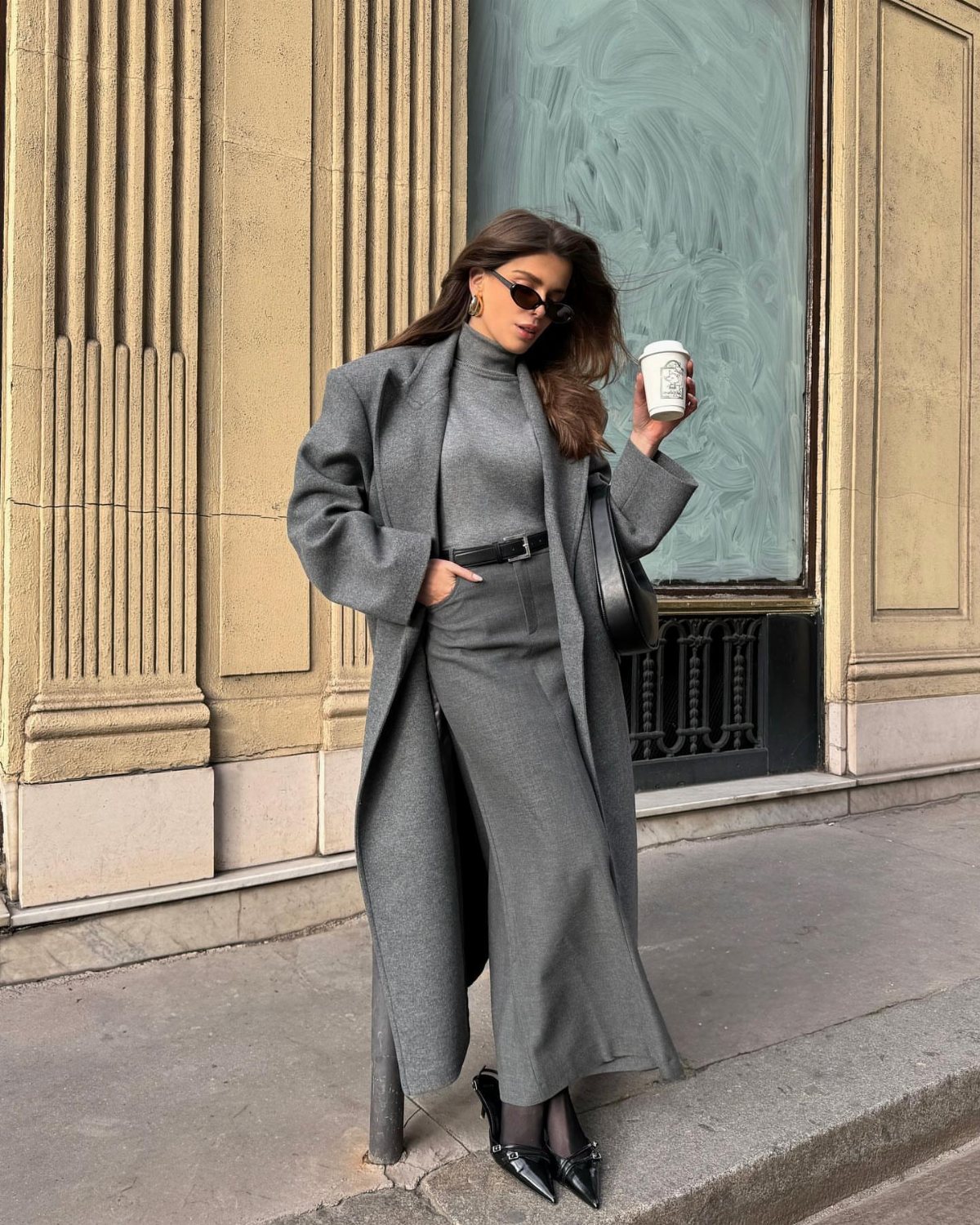 Woman in a monochrome grey long coat and turtleneck dress standing on a city sidewalk.