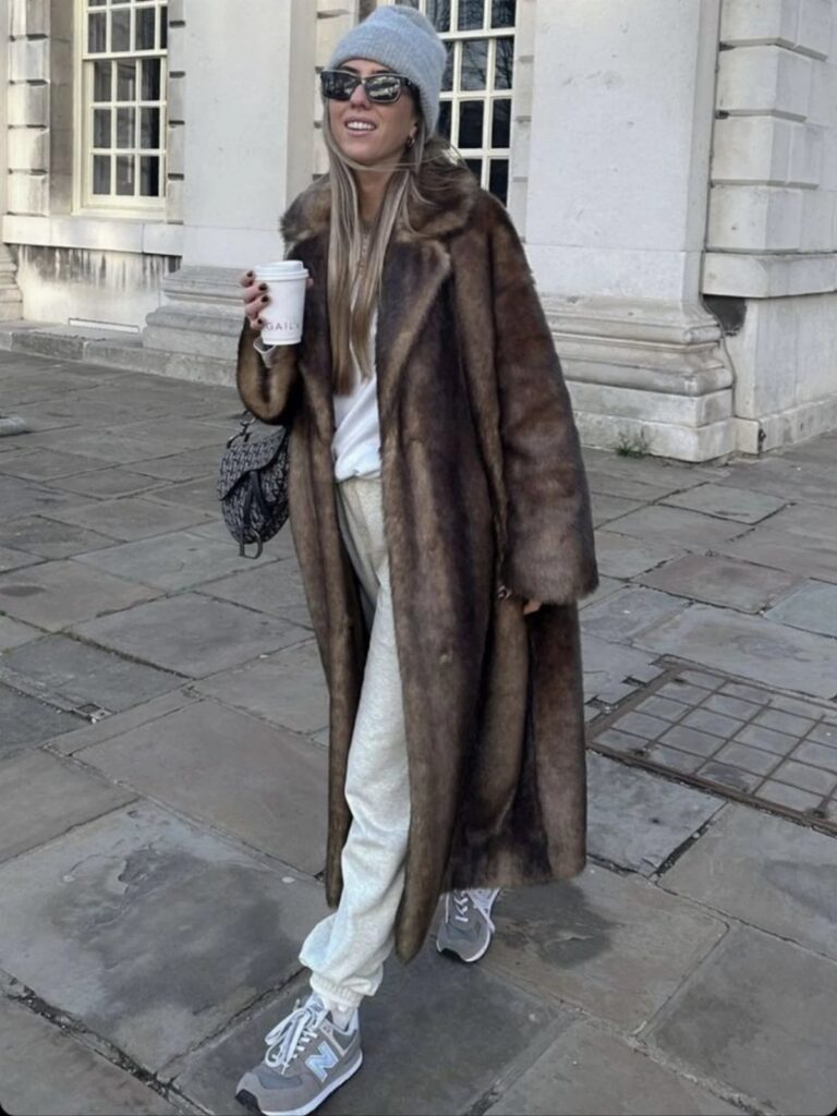 A woman walking outdoors wearing a long brown fur coat, grey sweatpants, and a beanie.