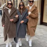 Three women walking down a city street wearing long teddy coats in brown, grey, and tan.