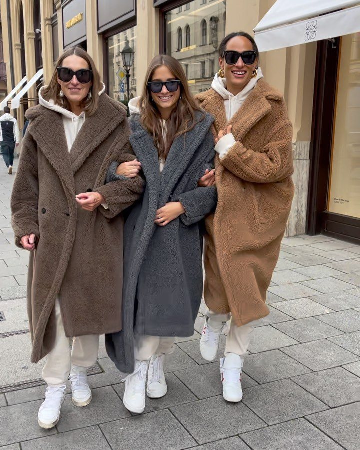 Three women walking down a city street wearing long teddy coats in brown, grey, and tan.