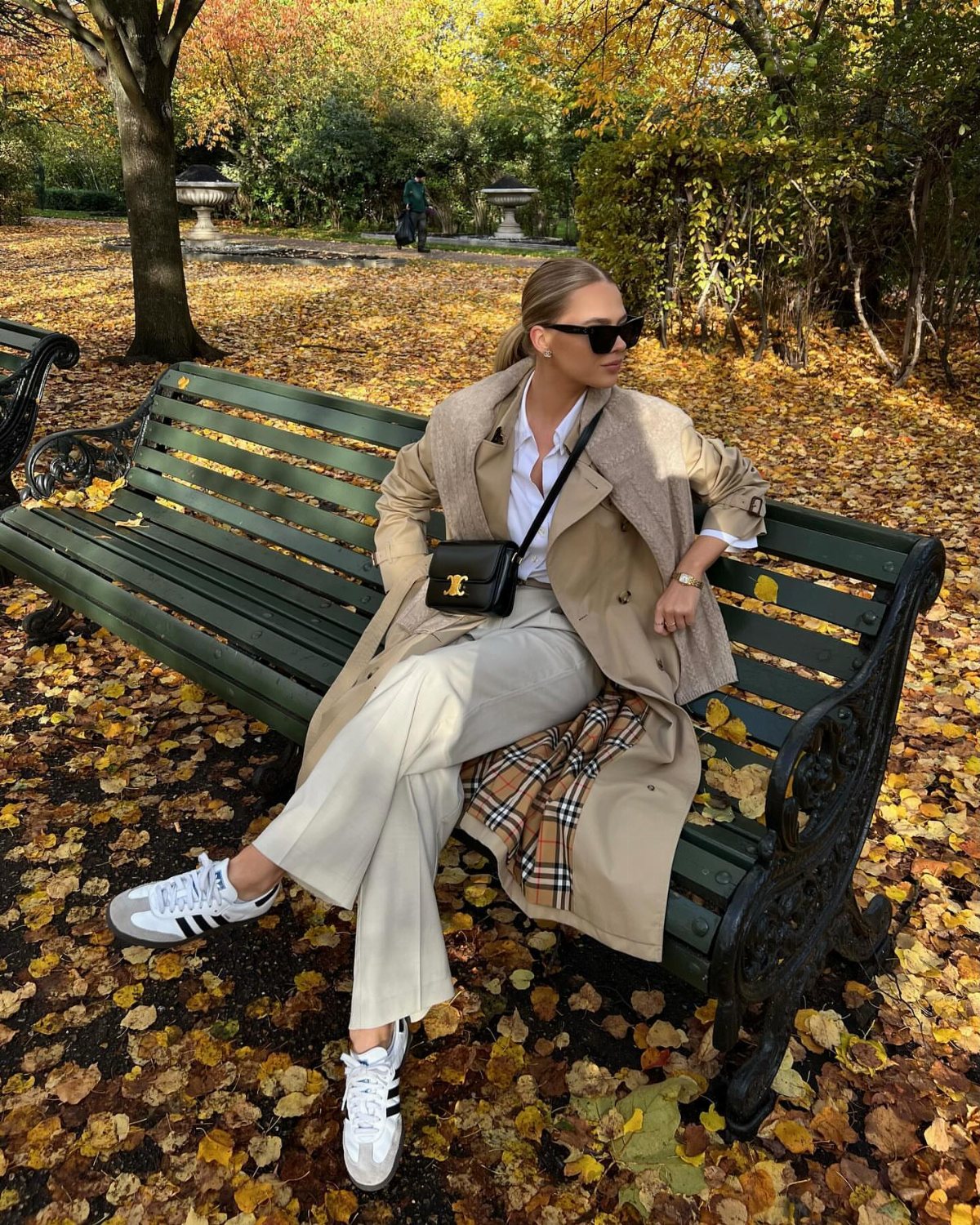 Woman in a beige trench coat and cream trousers sitting on a park bench wearing white sneakers.