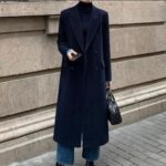 Woman in a long navy wool coat, black turtleneck, and blue jeans standing outdoors.