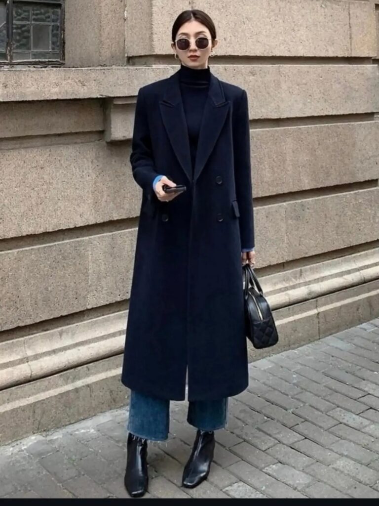 Woman in a long navy wool coat, black turtleneck, and blue jeans standing outdoors.