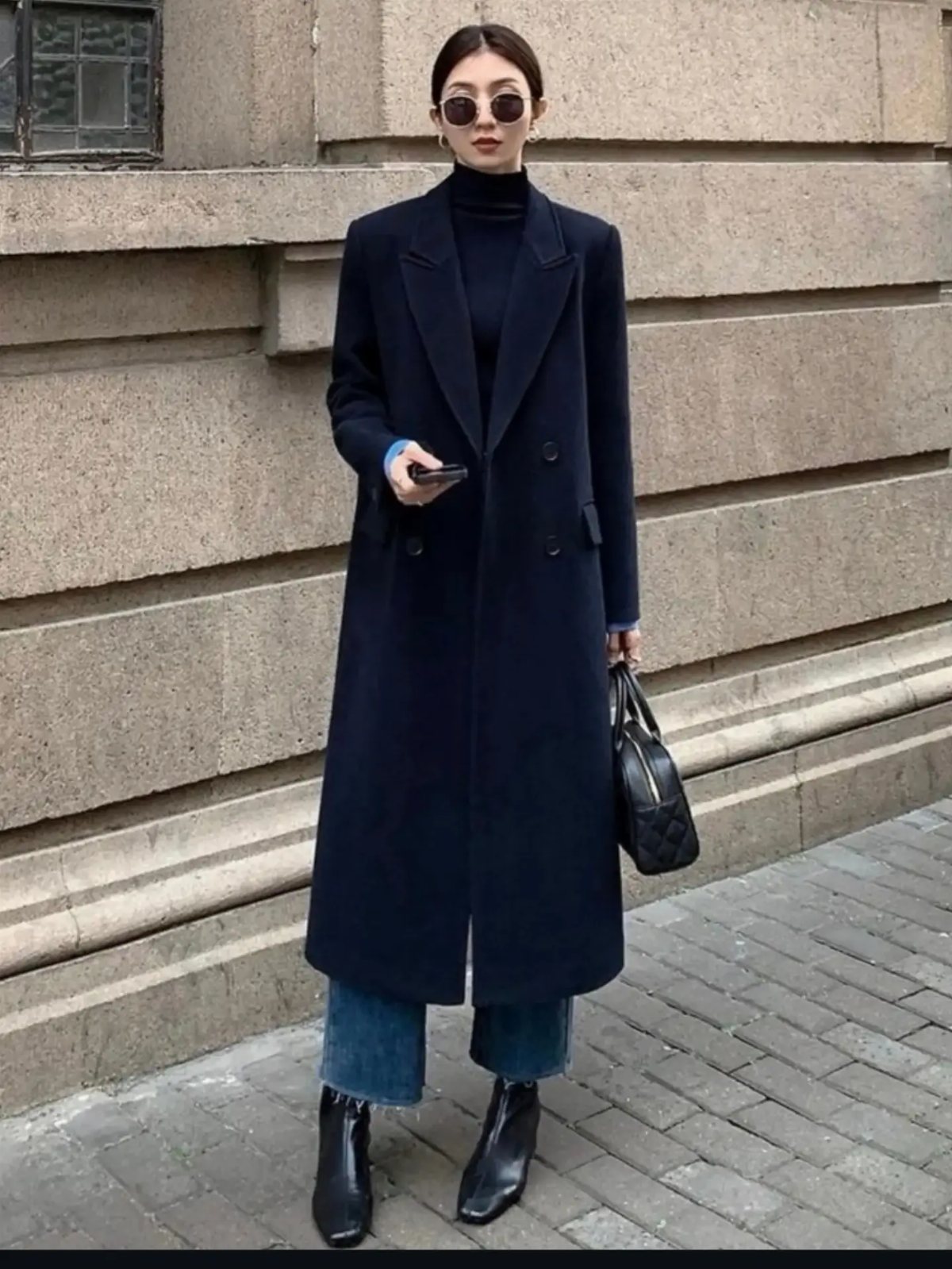 Woman in a long navy wool coat, black turtleneck, and blue jeans standing outdoors.