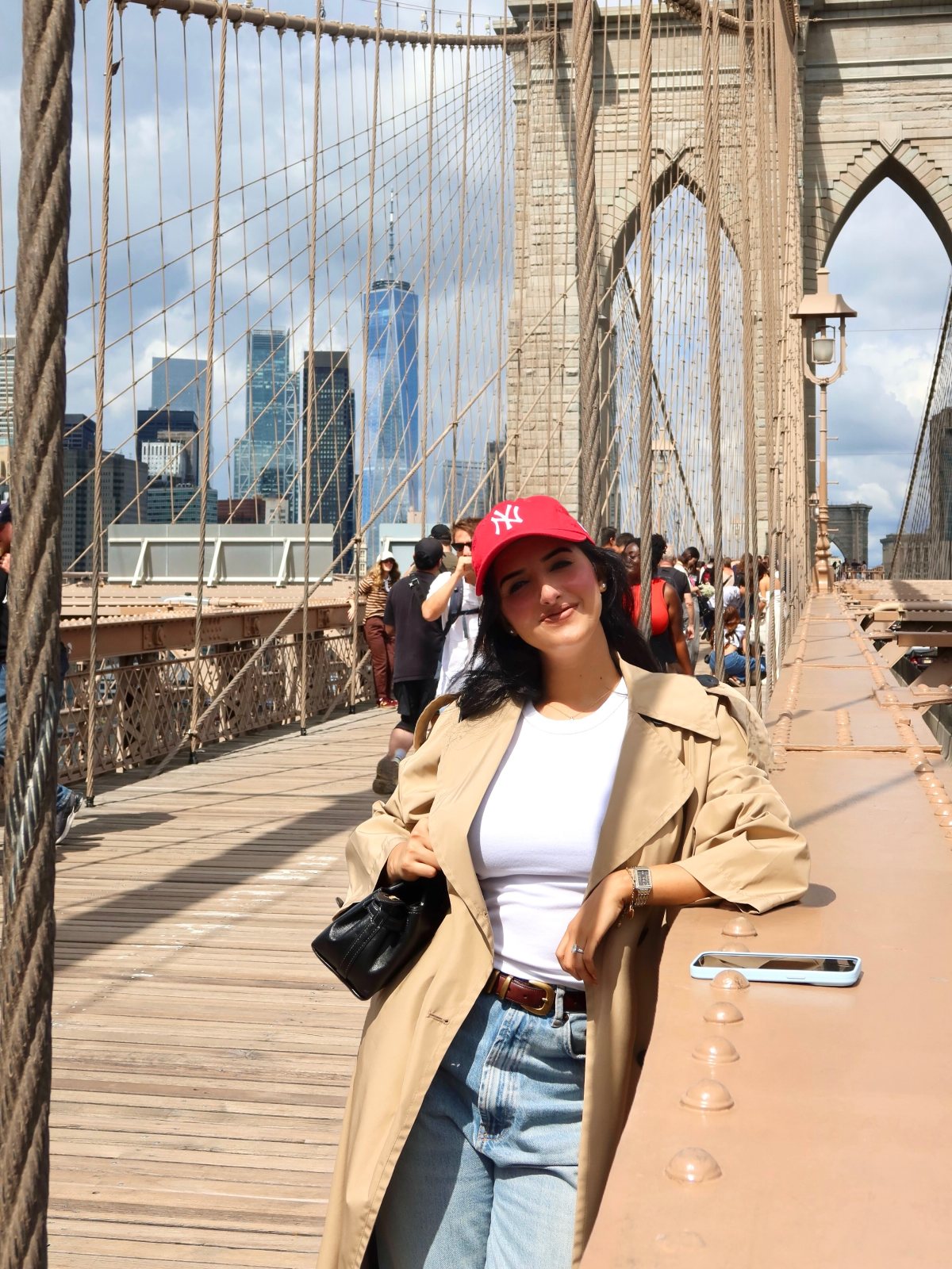 A woman in a tan trench coat and red Yankees cap posing on the Brooklyn Bridge.