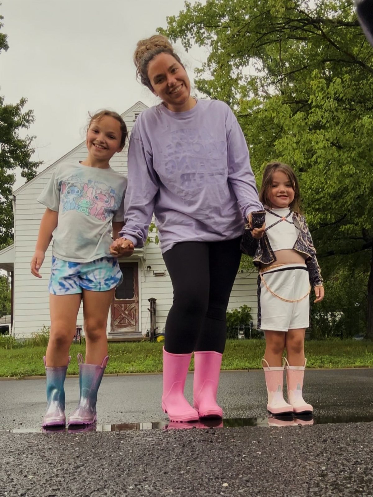 Three smiling people standing on wet pavement wearing colorful rubber rain boots.