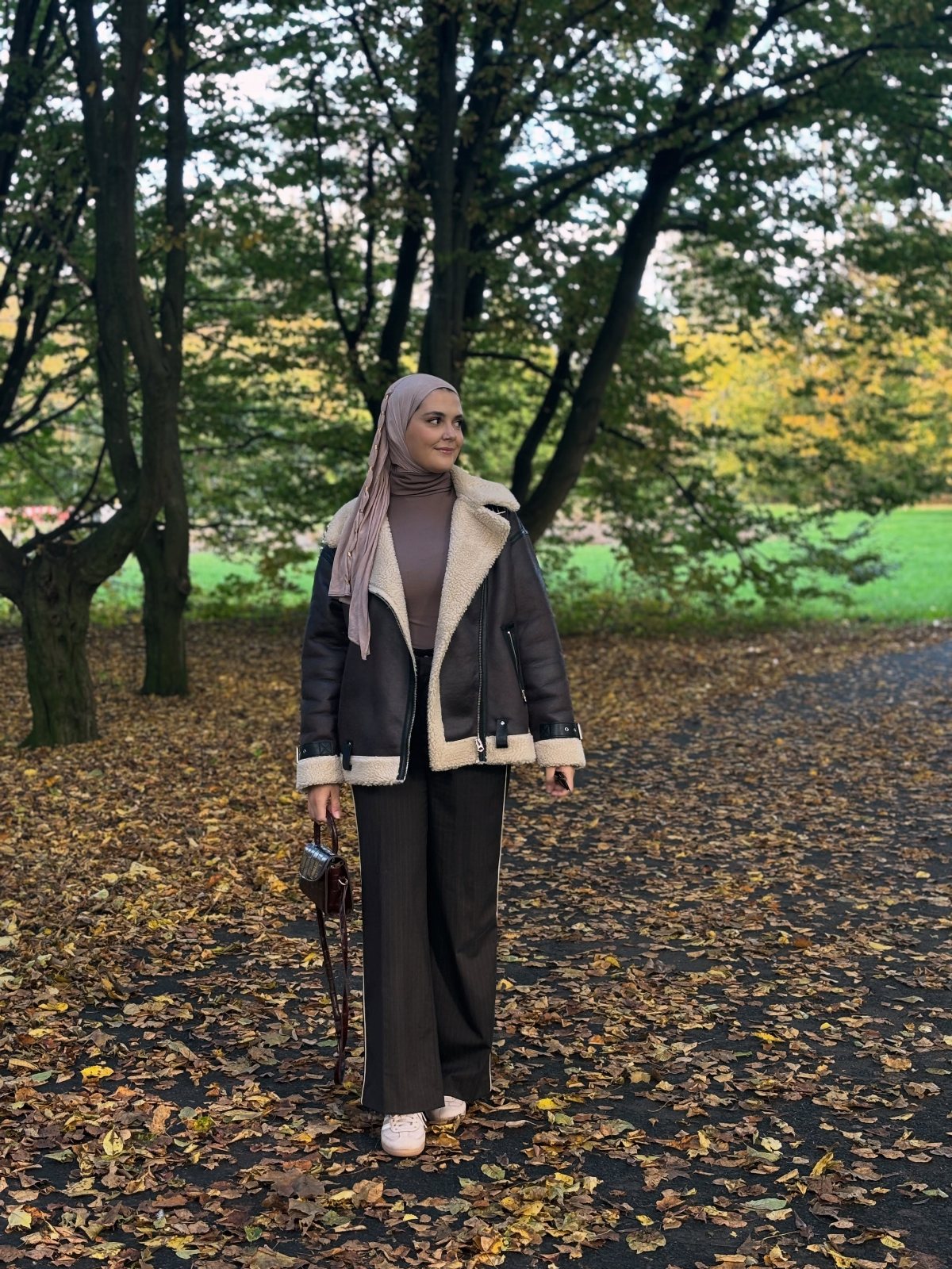 Woman in a shearling jacket and hijab standing in a park with autumn leaves.