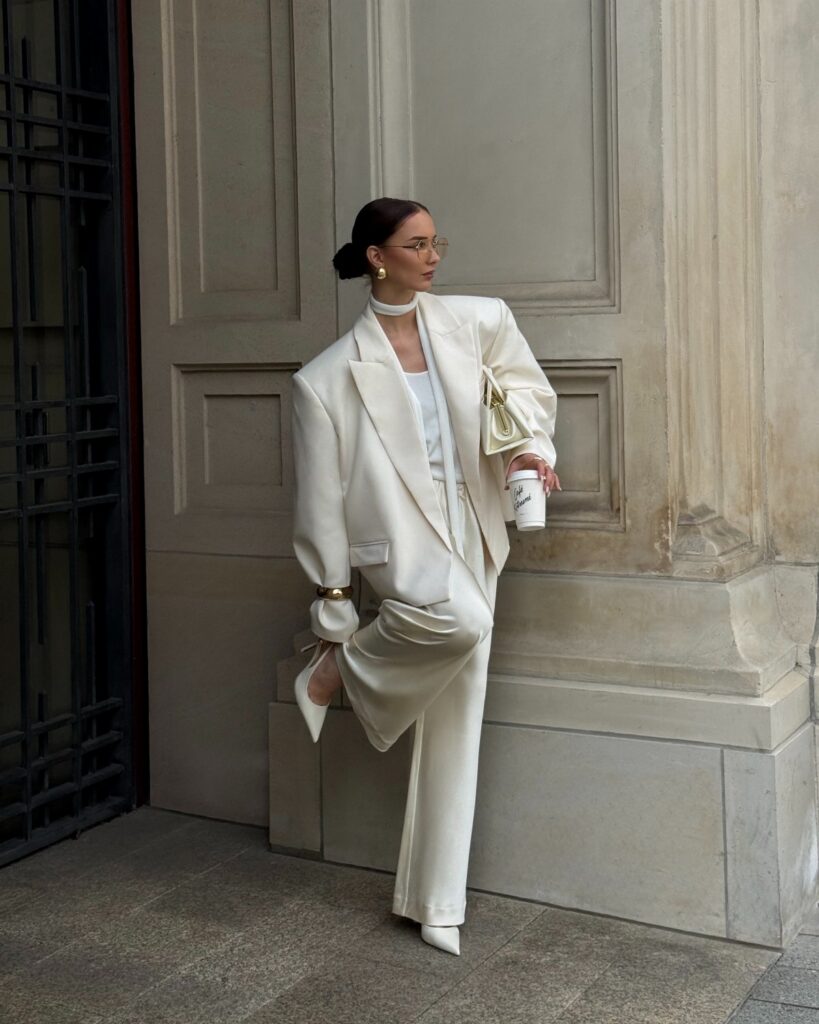 Woman wearing a cream monochromatic suit and white blazer standing against a stone wall.