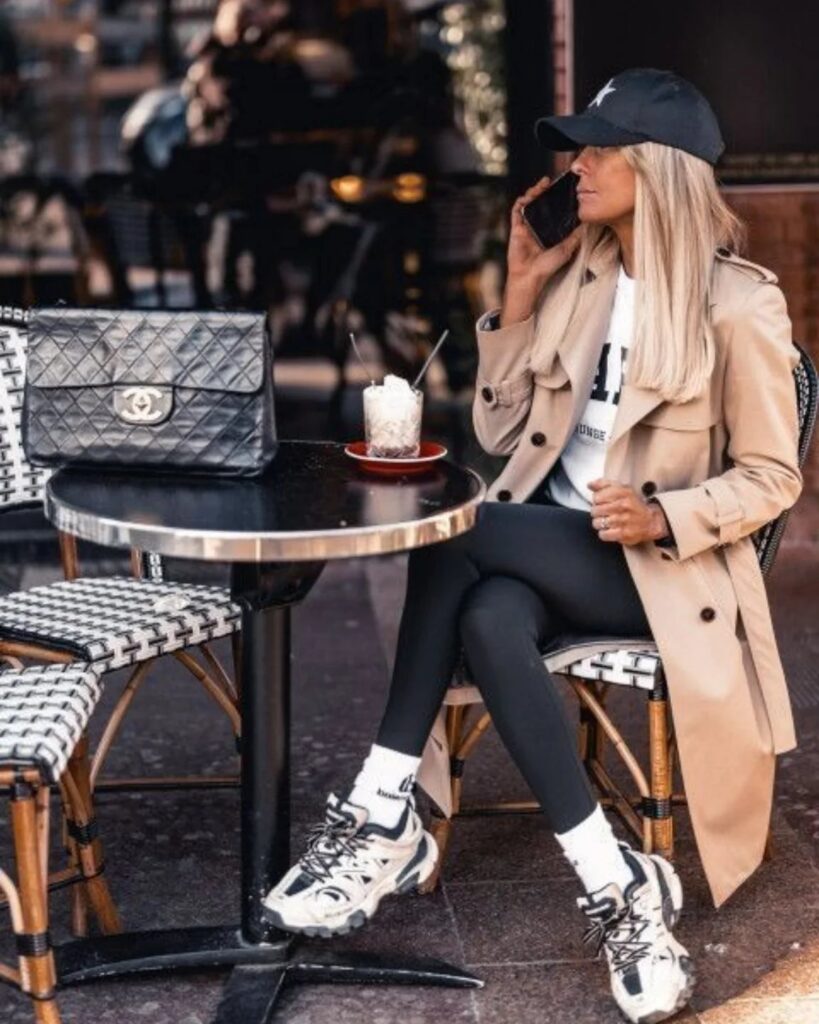 A woman in a tan trench coat and black leggings wearing chunky white sneakers while sitting at a cafe.